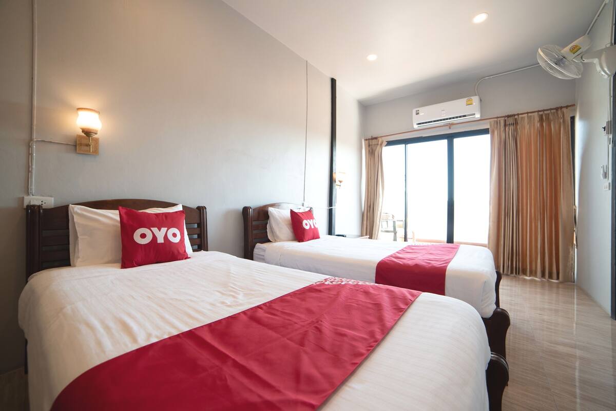 Une chambre d’hôtel avec deux lits, chacun recouvert d’une courtepointe rouge et blanche et agrémenté d’un oreiller orné du logo « OYO ». La chambre est équipée d’un ventilateur de plafond, d’un climatiseur et d’une fenêtre avec des rideaux.