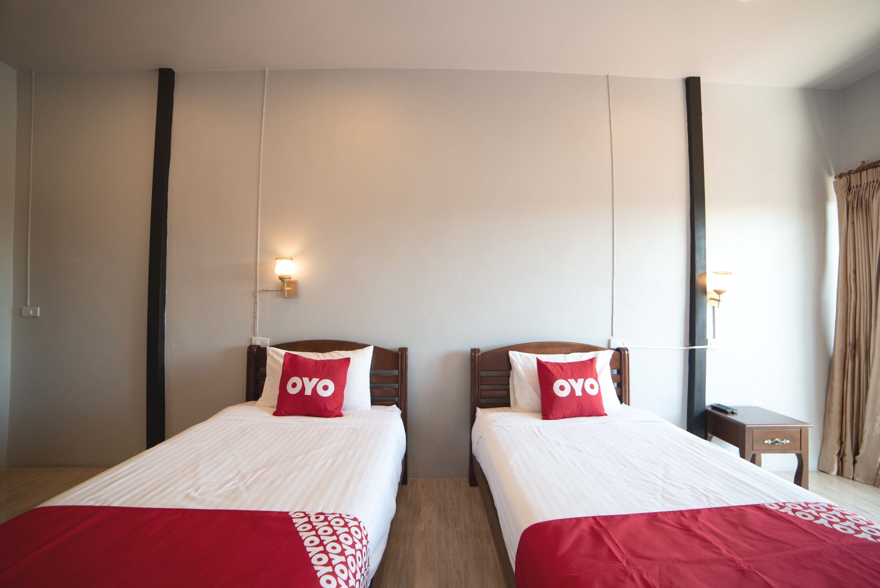 Habitación de hotel con dos camas, cada una con colcha roja y blanca, y un cojín con la palabra 'OYO'. Hay una mesita de noche con una lámpara y una pequeña mesa.