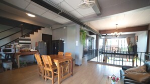 A spacious living area with a wooden dining table, a black leather sofa, and a glass coffee table. There is a kitchen area with a microwave and a refrigerator. A staircase leads to an upper level with a balcony. - OYO 465 Krung Kao Traveller Lodge (Ayutthaya)