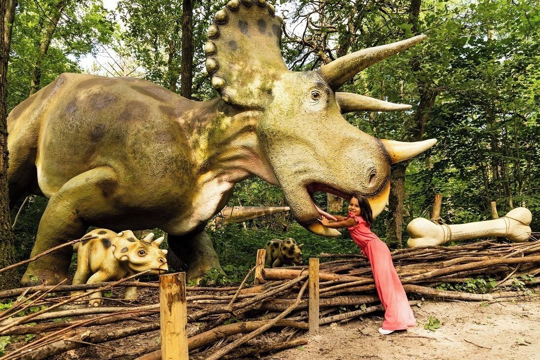 Une grande statue de dinosaure dans une zone boisée, où un enfant interagit avec elle.