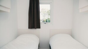 Een kleine hotelkamer met één ruimte, twee eenpersoonsbedden, een raam met gordijn en uitzicht op bomen.