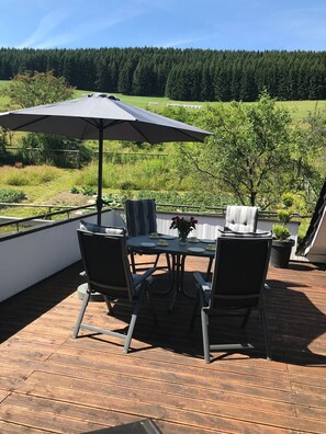 Une terrasse avec une table et des chaises, un grand parasol et une vue sur une zone boisée.