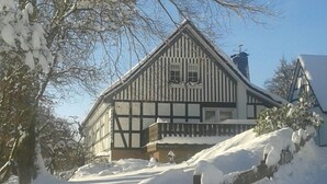 Une maison traditionnelle à colombages, dotée d’un toit à forte pente, entourée d’un paysage enneigé et d’arbres.