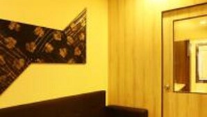 A room with a dark-colored sofa against a yellow wall, a wooden door, and a patterned wall hanging. - Balaji Residency (Chennai)