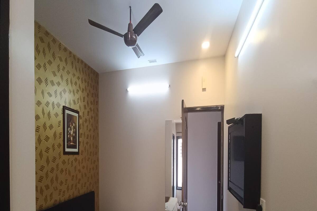 Habitación de hotel con cama, ventilador de techo, televisión y puerta que da a un baño.