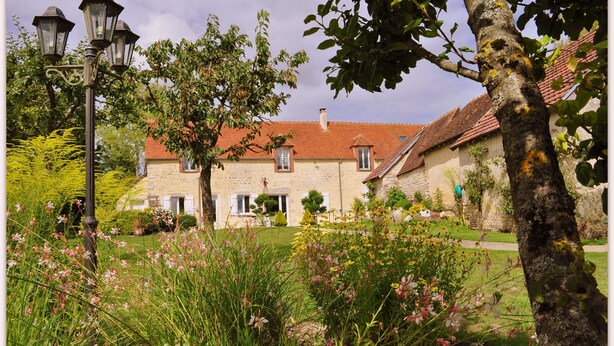 A stone building with a red-tiled roof, surrounded by lush greenery and flowering plants. - Le Bourg d'Aunou (Aunou-sur-Orne)