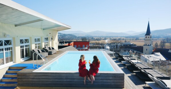 A rooftop pool with two people in red robes, a view of a church, and a mountainous landscape in the background. - Hotel Randsbergerhof (Cham)
