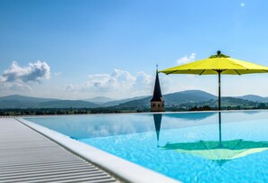 A swimming pool with a yellow umbrella, a clock tower, and a mountainous landscape in the background.