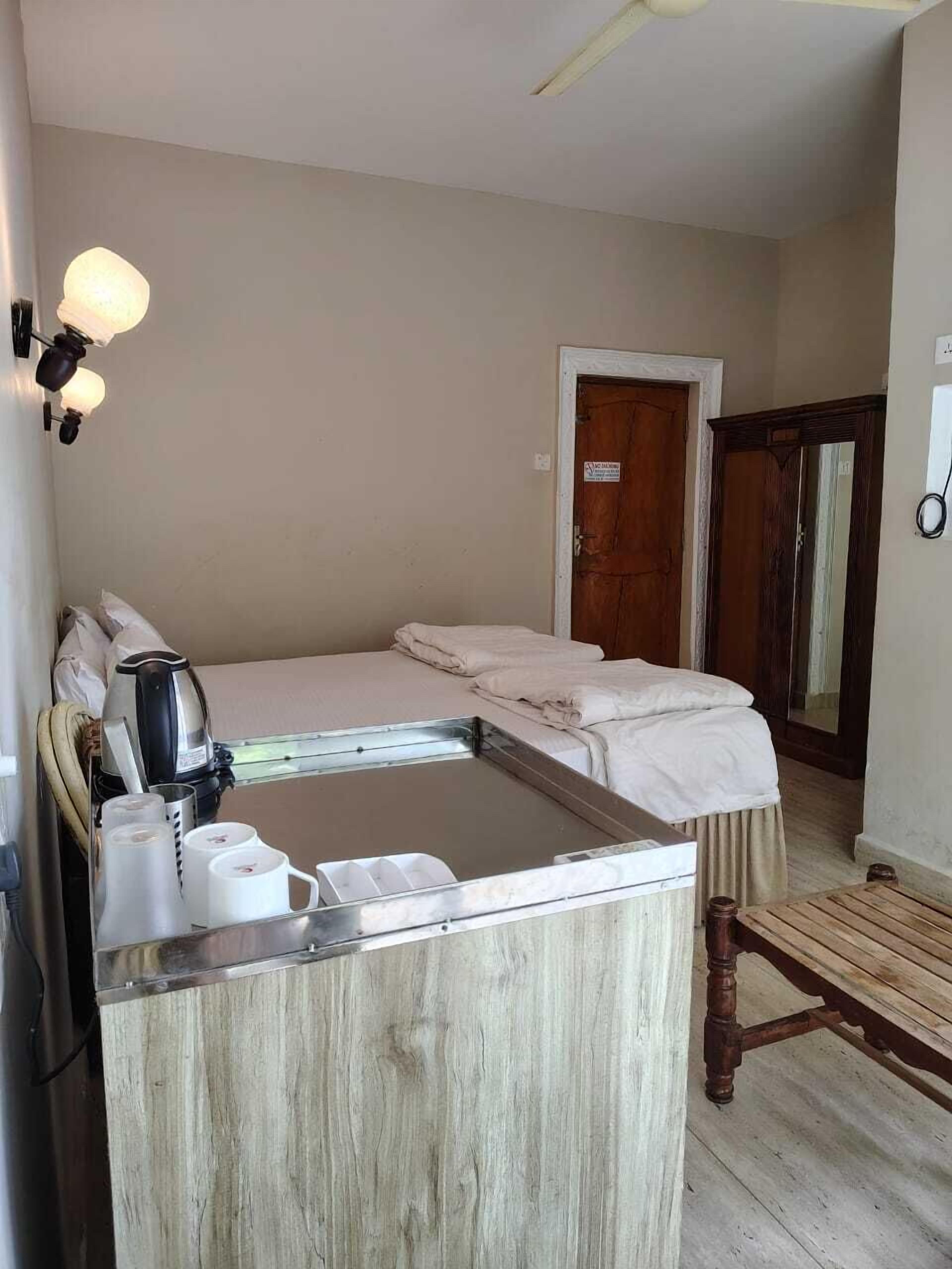Una habitación con cama, una mesita con tetera y tazas, un banco de madera y una puerta.