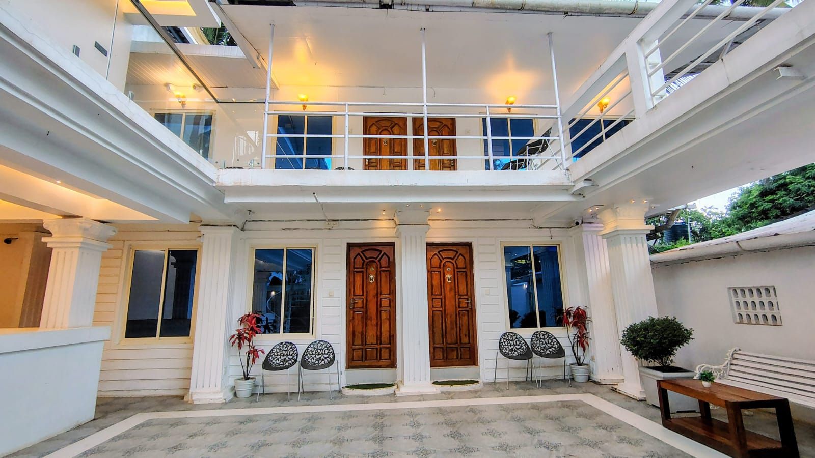 A multi-level building with a central entrance featuring two wooden doors, a balcony with railings, and a seating area with chairs and a bench.