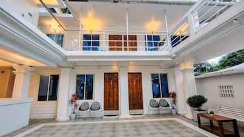A multi-level building with a central entrance featuring two wooden doors, a balcony with railings, and a seating area with chairs and a bench.