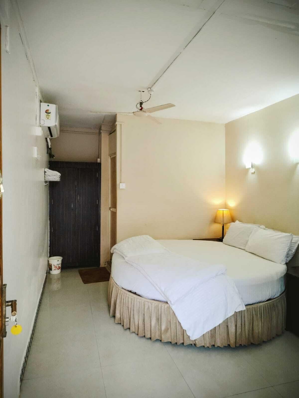A small, single-bed room with a ceiling fan, a bedside lamp, and a door leading to a dark room.