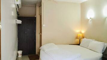 A small, single-bed room with a ceiling fan, a bedside lamp, and a door leading to a dark room.