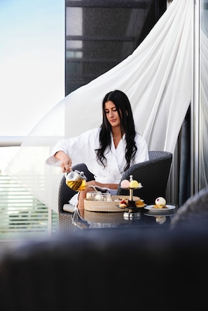 Une personne est assise à une table où se trouvent divers aliments, notamment une théière et un plateau de pâtisseries. La scène se déroule dans un cadre extérieur moderne, avec un auvent blanc et une rambarde en verre.