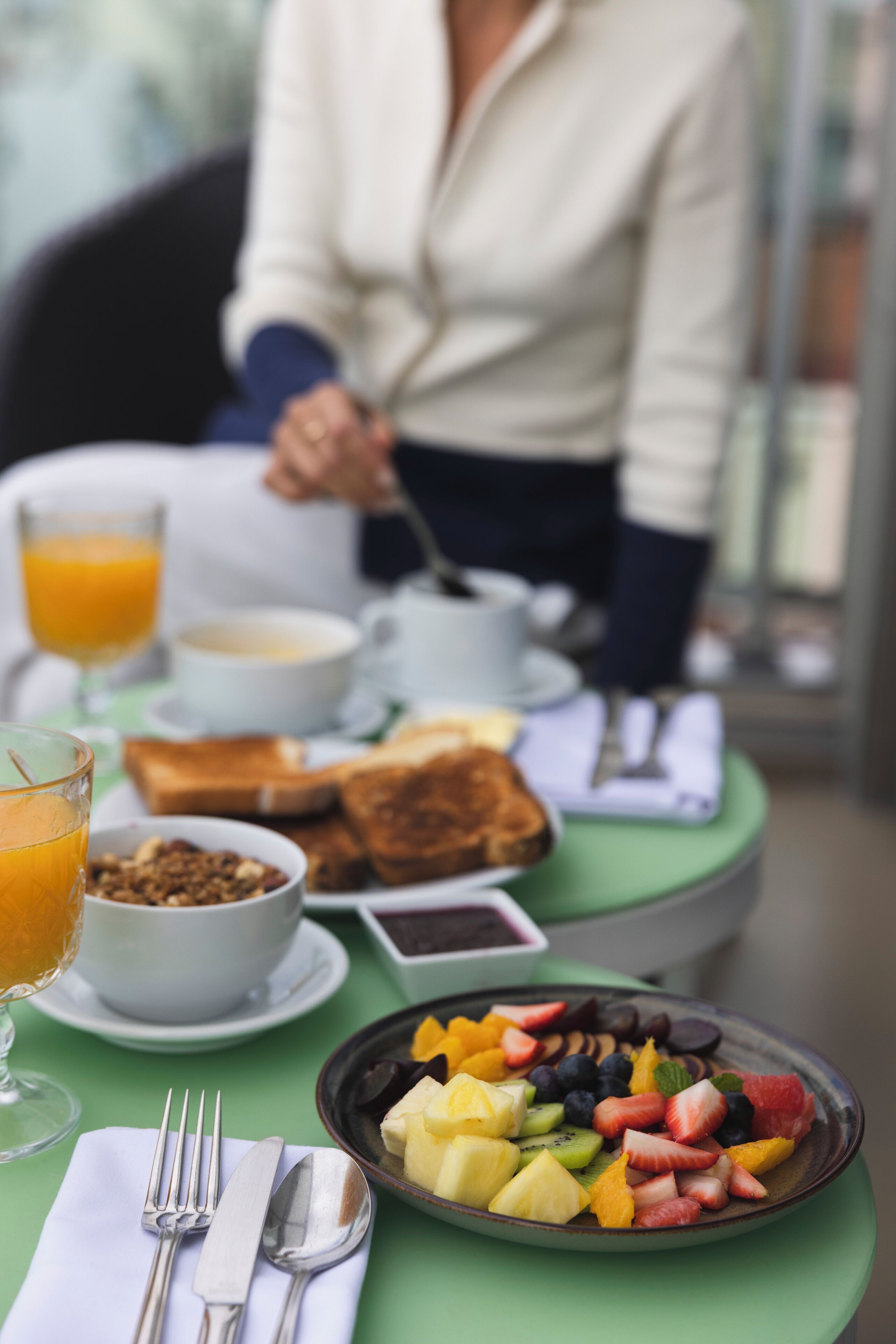 A breakfast table with a plate of fruit, a bowl of cereal, a glass of orange juice, and a cup of coffee.