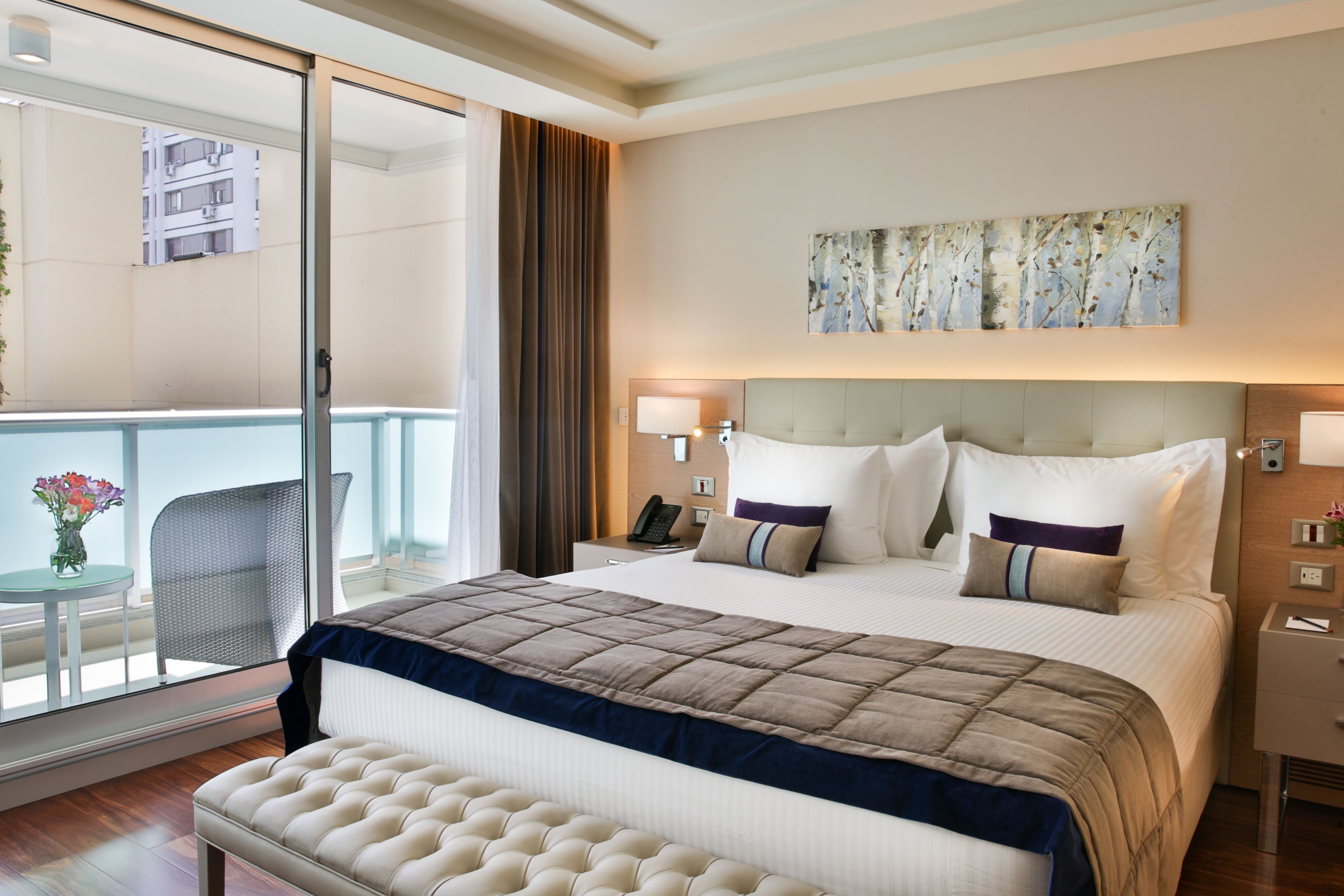 A hotel room with a large bed, a bedside table, a chair, a balcony with a view of buildings, and a painting on the wall.