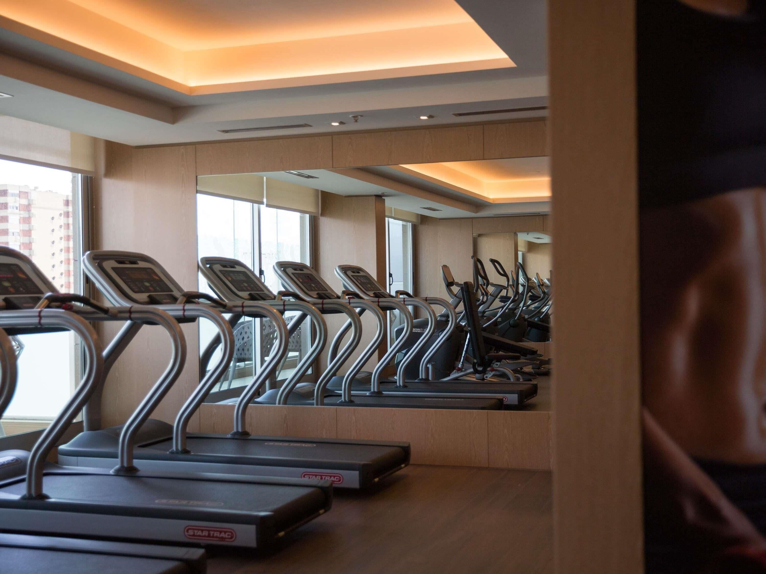 Una fila de cintas de correr en un gimnasio de diseño moderno, con amplios ventanales y un espejo grande.