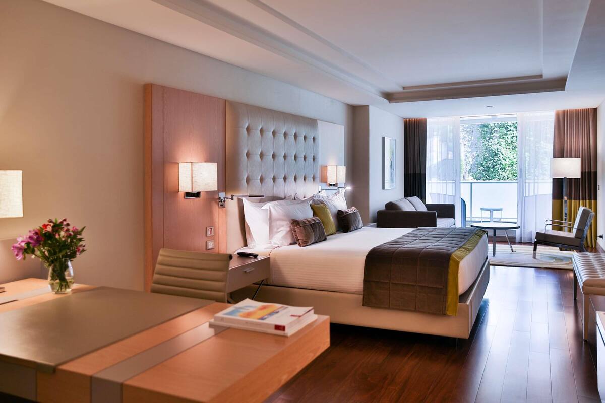 Hotelkamer met een groot bed, een bureau, een stoel en een klein tafeltje. Er is een raam met gordijnen en uitzicht op bomen.