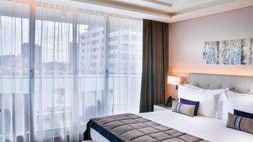 A hotel room with a large bed, a bench, a bedside table with a lamp, and a view of buildings through sheer curtains.