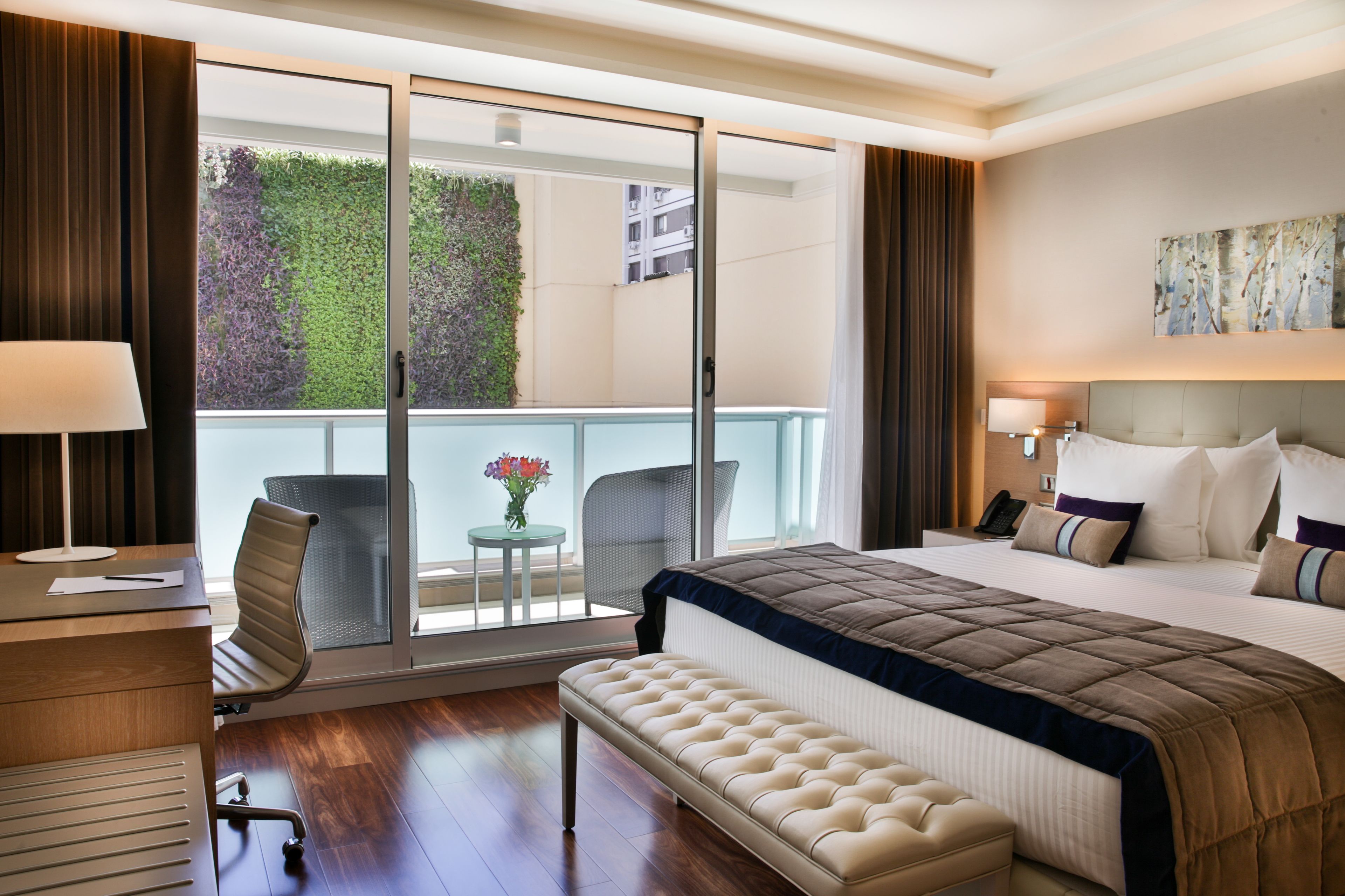 A hotel room with a large bed, a desk with a chair, a bench, a balcony with a chair and table, and a view of greenery outside.