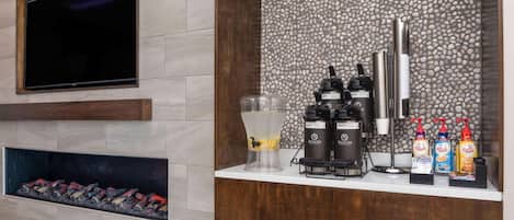 Eine moderne Hotelhalle mit einem an der Wand befestigten Flachbildfernseher, einem Kamin mit Holzscheiten, einer Kaffeestation mit mehreren Kaffeemaschinen und einer Wandverkleidung aus kleinen Kieselsteinen.