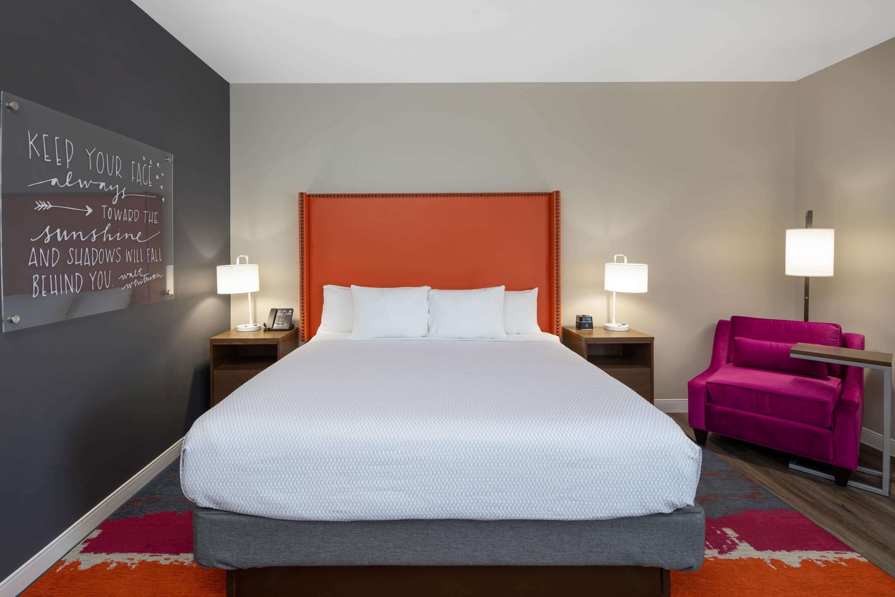 Habitación de hotel con una cama grande, dos mesitas de noche con lámparas, un cabecero naranja brillante, una silla rosa y una frase motivacional en la pared.