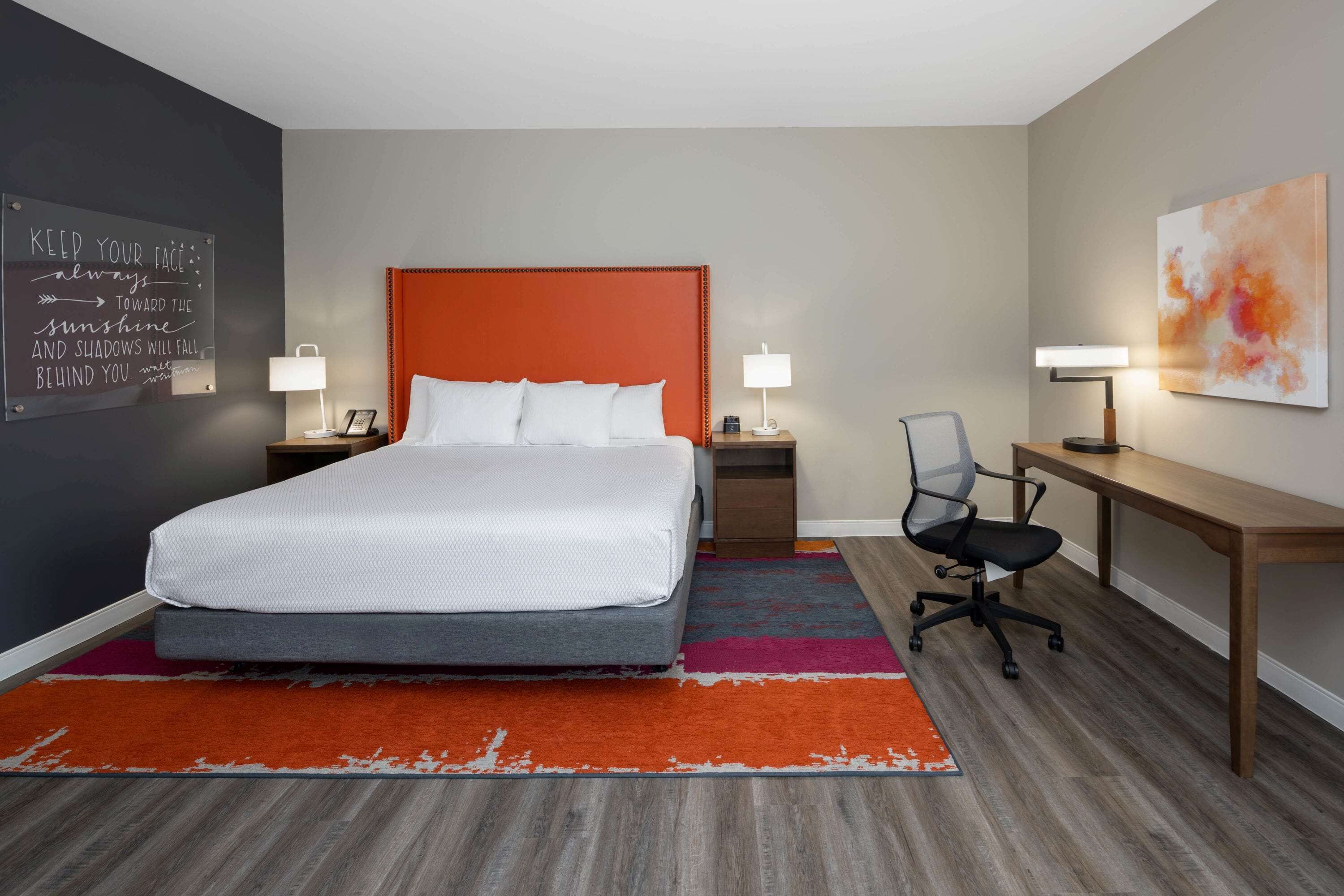 Habitación de hotel con cama, mesitas de noche, escritorio, silla y una cita motivacional en la pared.