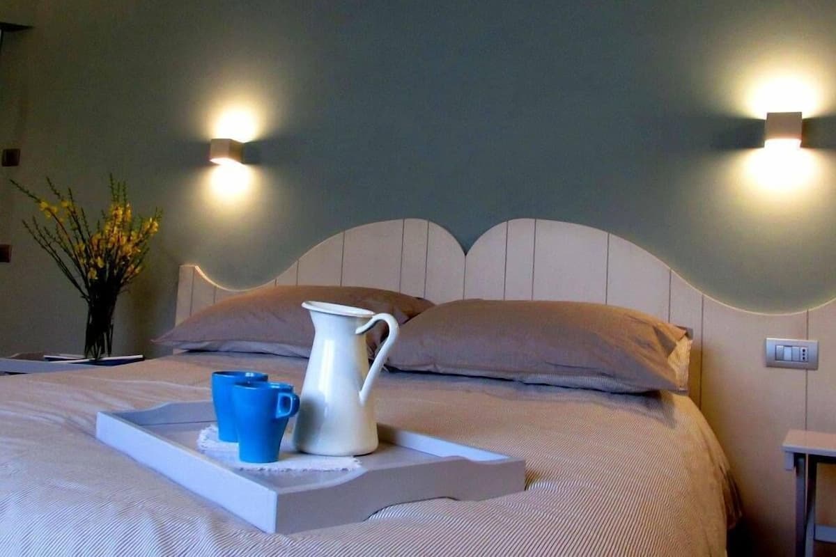 A neatly made bed with a white headboard, two pillows, a white pitcher, and two blue cups on a tray.