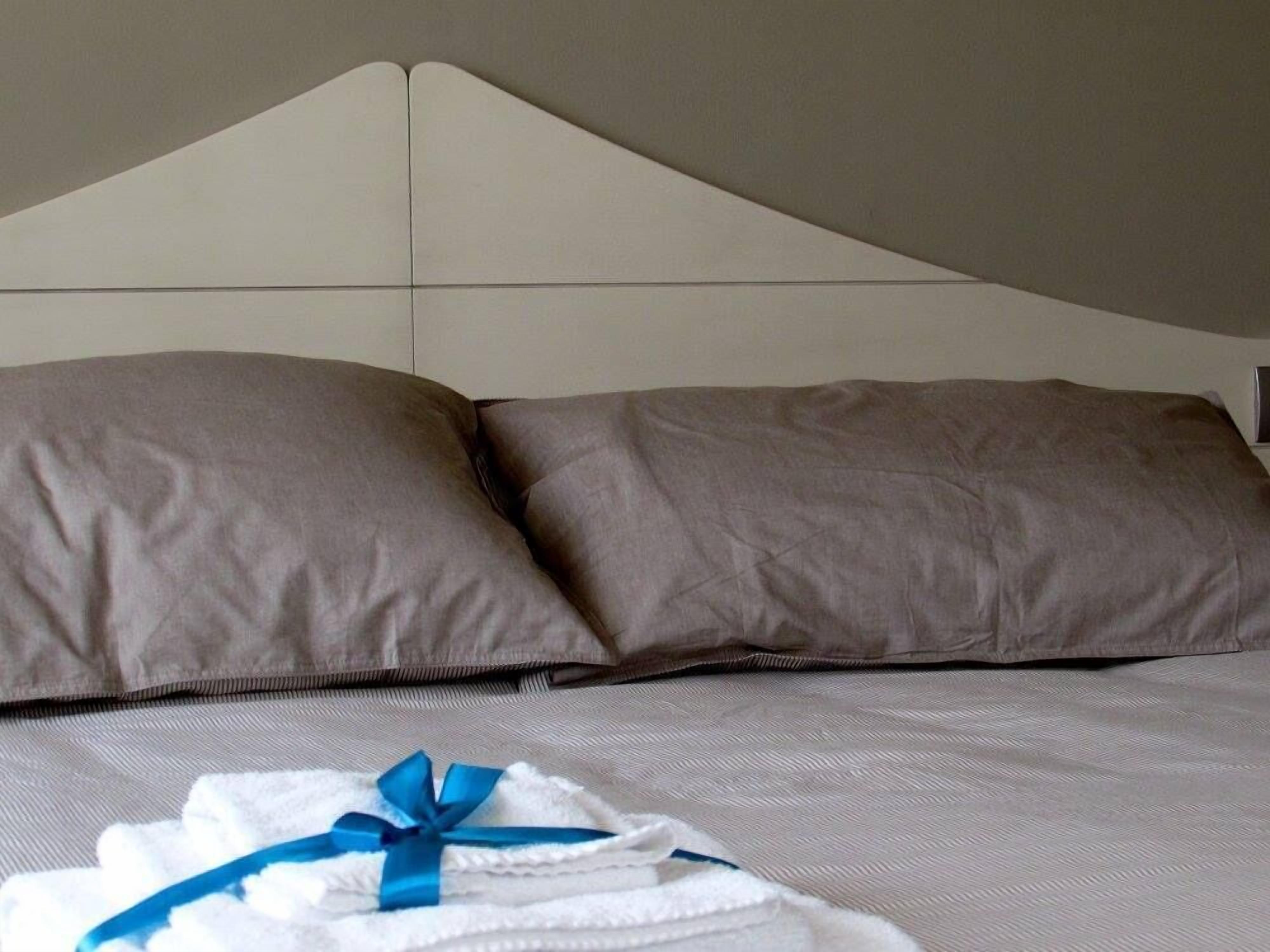 Ein ordentlich bezogenes Bett mit zwei Kissen und einem gefalteten weißen Handtuch mit blauer Schleife.