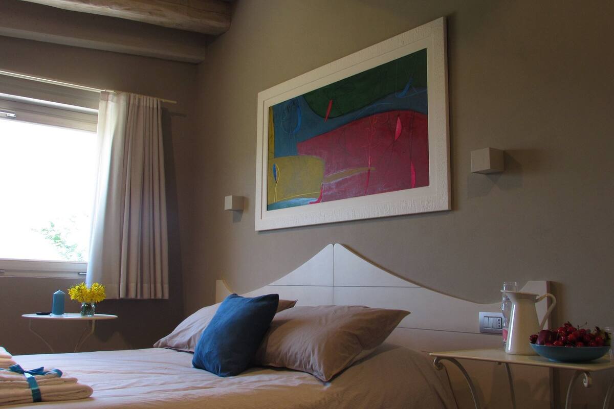 A hotel room with a bed, a painting on the wall, a small table with a vase of flowers, and a pitcher of fruit on a side table.
