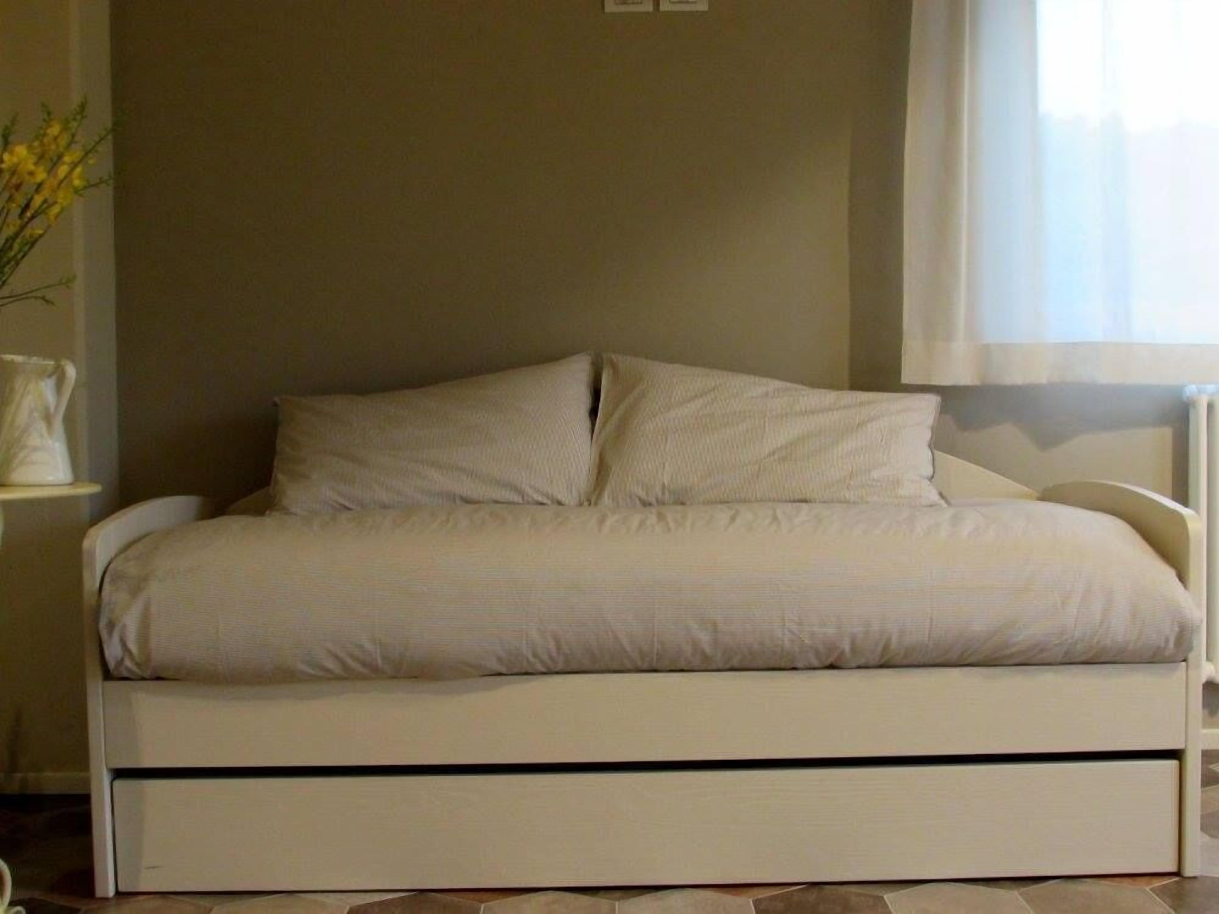Un lit avec un cadre blanc et un matelas, un vase avec des fleurs jaunes et une fenêtre avec des rideaux.