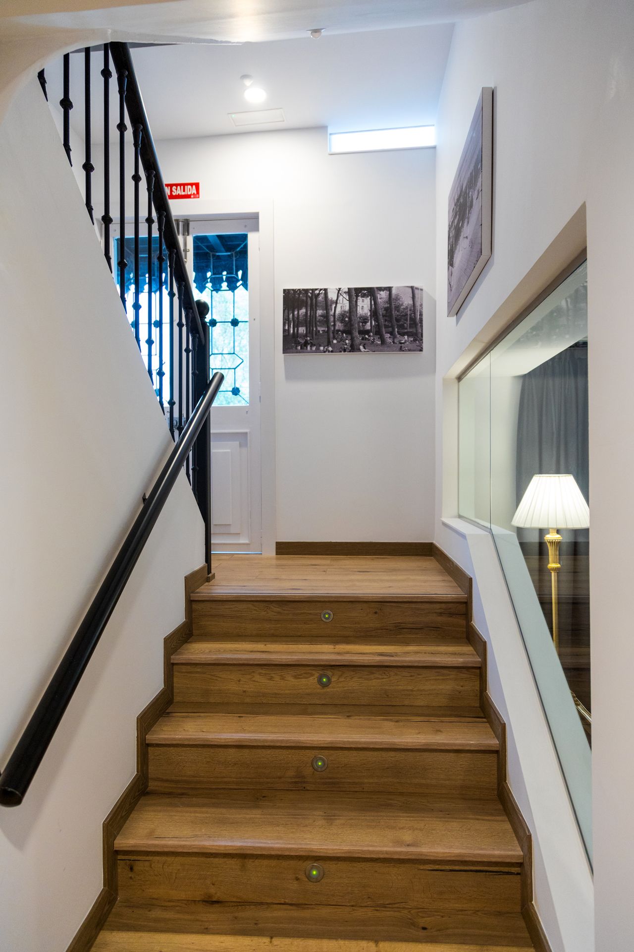 A wooden staircase with black balusters, a handrail, and a red exit sign.