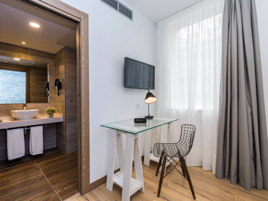 A modern bathroom with a large mirror, a sink, and a shower. There is a glass table with a chair and a TV mounted on the wall.