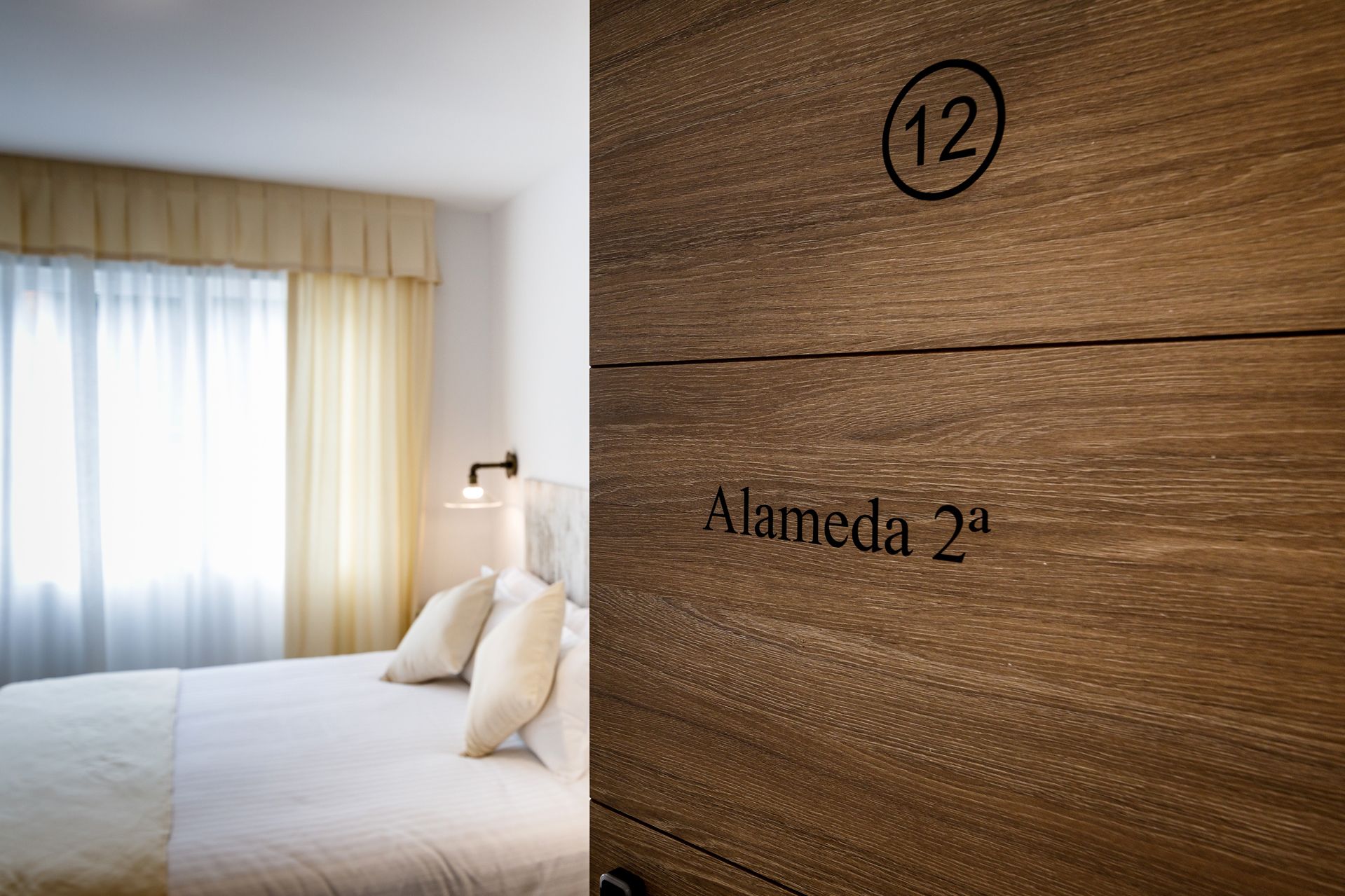 A hotel room with a bed, a nightstand, and a wooden wall with the number 12 and the name "Alameda 2a".