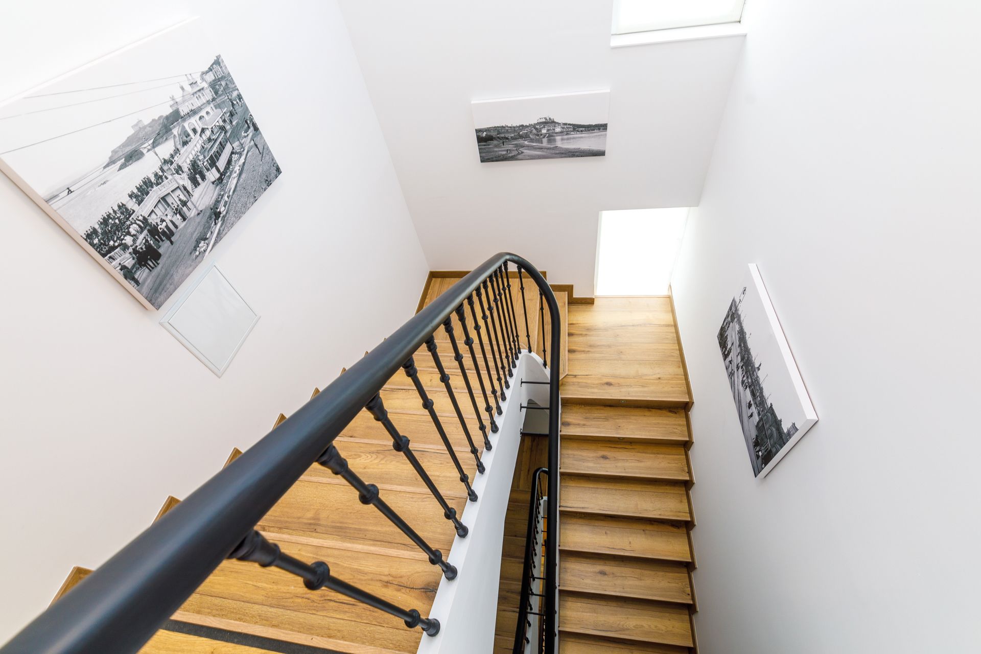 A staircase with black railings and wooden steps, leading to a landing with three black-and-white photographs on the walls.