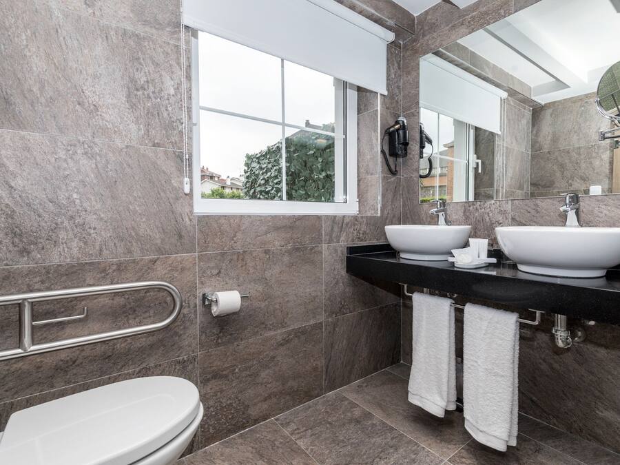 Un baño moderno con inodoro, dos lavamanos, pasamanos, ventana y espejo.