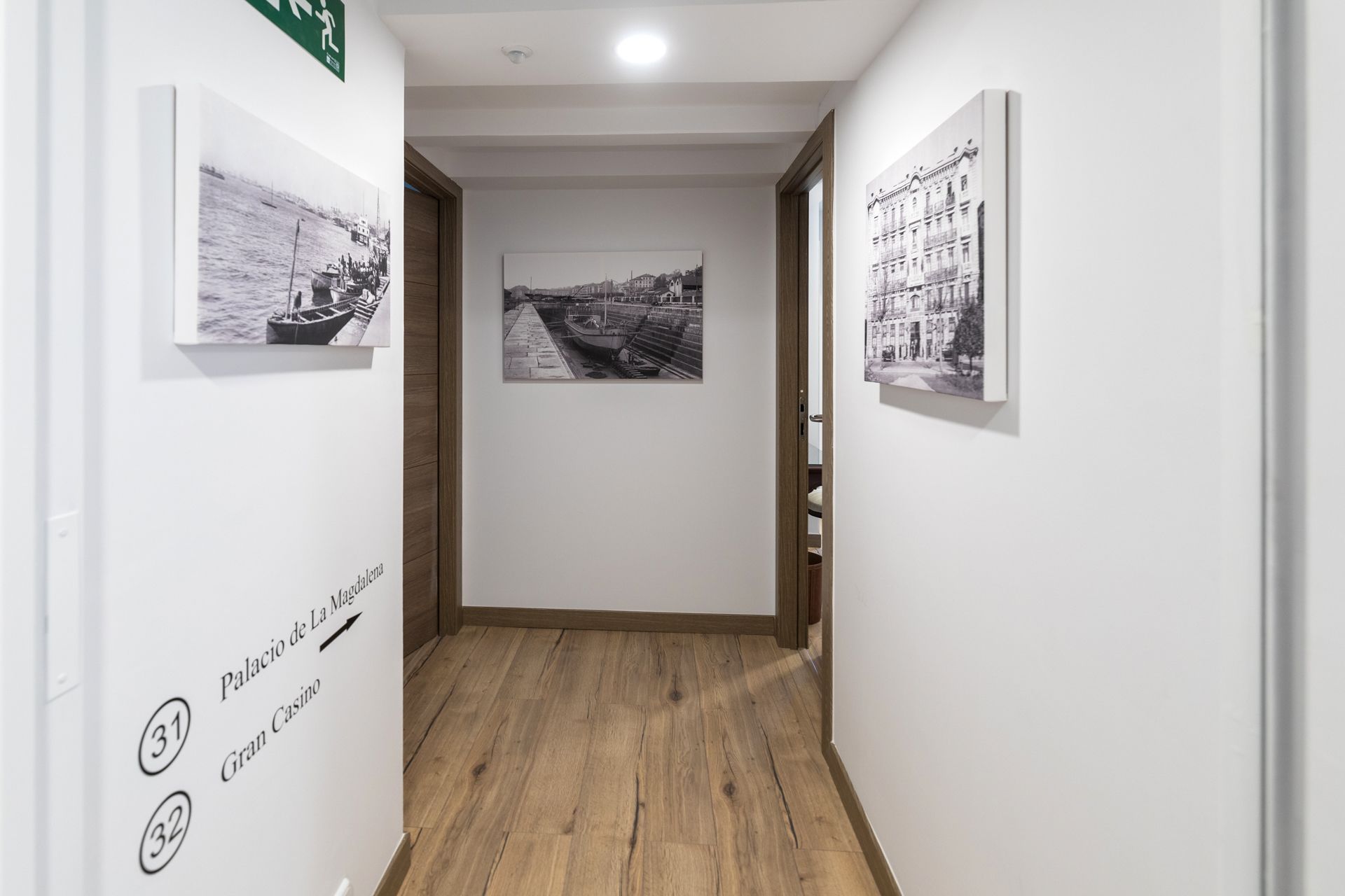 Un pasillo con suelo de madera, fotografías en blanco y negro en las paredes y una puerta que da a otra habitación.
