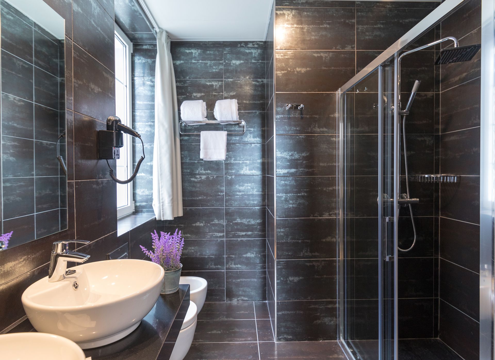 A modern bathroom with a large white sink, a glass shower enclosure, and dark tiled walls.