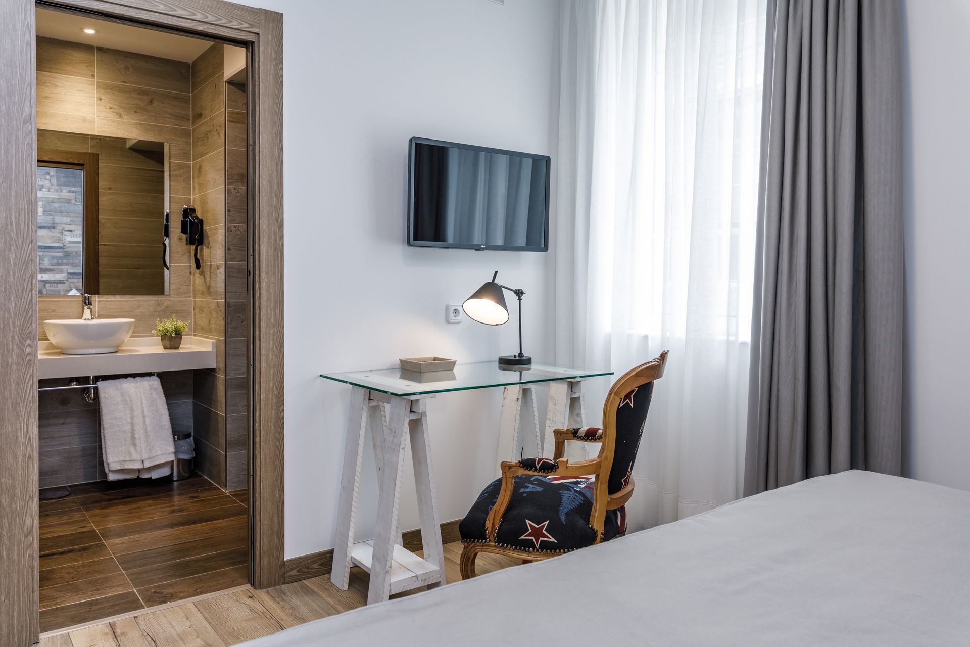Habitación de hotel con piso de madera, un escritorio de vidrio con lámpara, televisor, una silla y una cama grande.