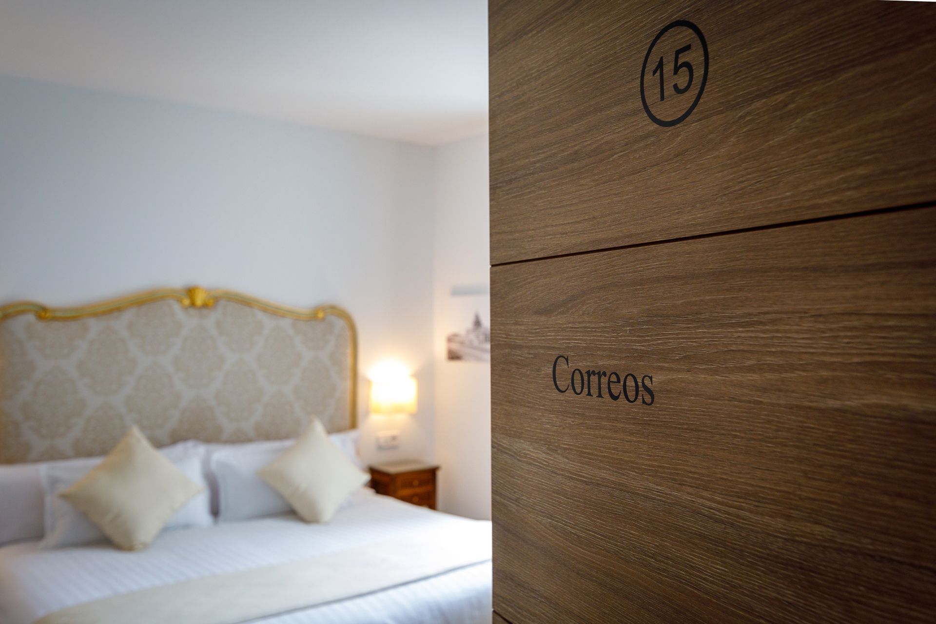 Une chambre d’hôtel avec un lit, des oreillers et un panneau en bois portant le numéro 15 et le mot « Correos ».