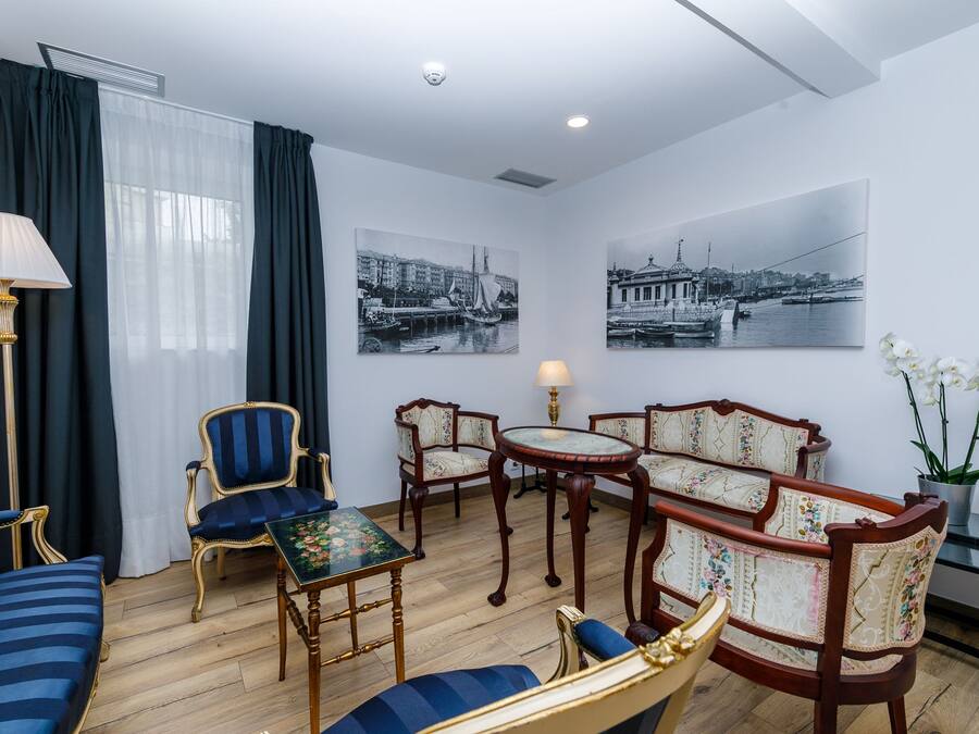 A hotel room with a wooden floor, a sofa, two armchairs, a small table, a lamp, and two black and white photographs on the wall.