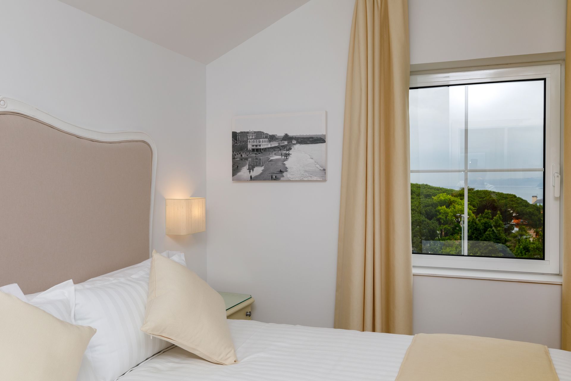 A hotel room with a bed, a nightstand, a lamp, a window with curtains, and a picture on the wall.