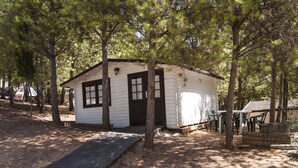 A small white cabin with a dark brown door and windows, surrounded by trees. - Camping ValdeArenas (Iznajar)
