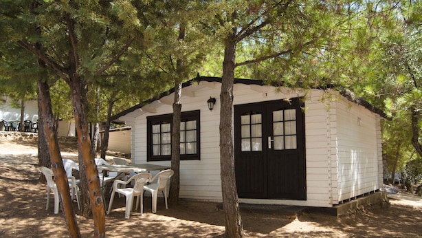 A small, single-story cabin with a white exterior and black trim, surrounded by trees. There is a table with chairs outside, and a window with a shutter. - Camping ValdeArenas (Iznajar)