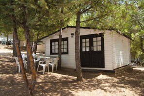 A small, single-story cabin with a white exterior and black trim, surrounded by trees. There is a table with chairs outside, and a window with a shutter. - Camping ValdeArenas (Iznajar)