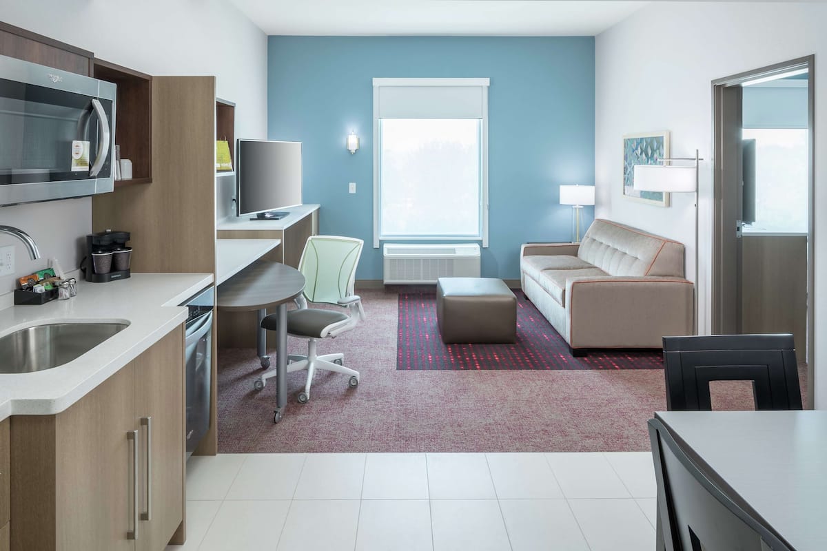 A modern hotel room with a kitchenette, a dining area, a sofa, and a large window providing natural light.