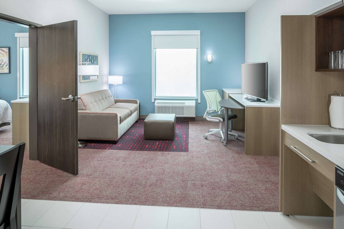 A modern hotel room with a sofa, a dining table, a TV, and a kitchenette.