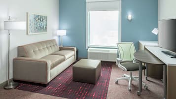 A hotel room with a sofa, ottoman, desk, chair, lamp, and artwork on the wall.