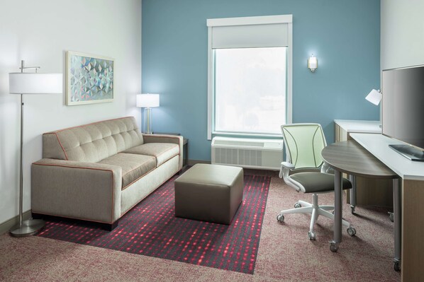 A hotel room with a sofa, ottoman, desk, chair, lamp, and artwork on the wall.