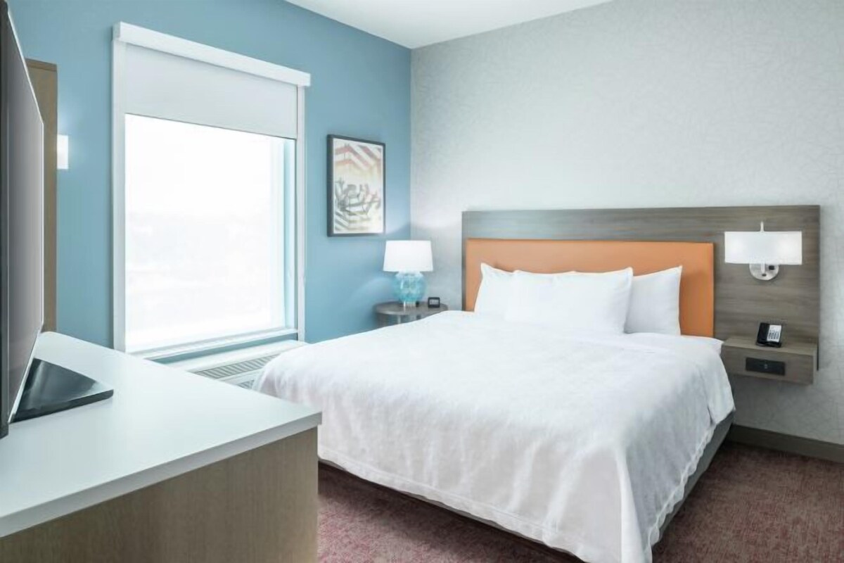 A hotel room with a bed, bedside lamps, a desk with a laptop, a window, and a wall-mounted artwork.
